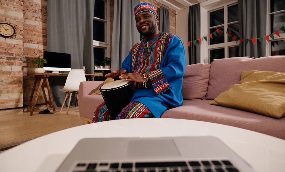 Smiling man in vibrant traditional African clothing playing a djembe drum indoors.